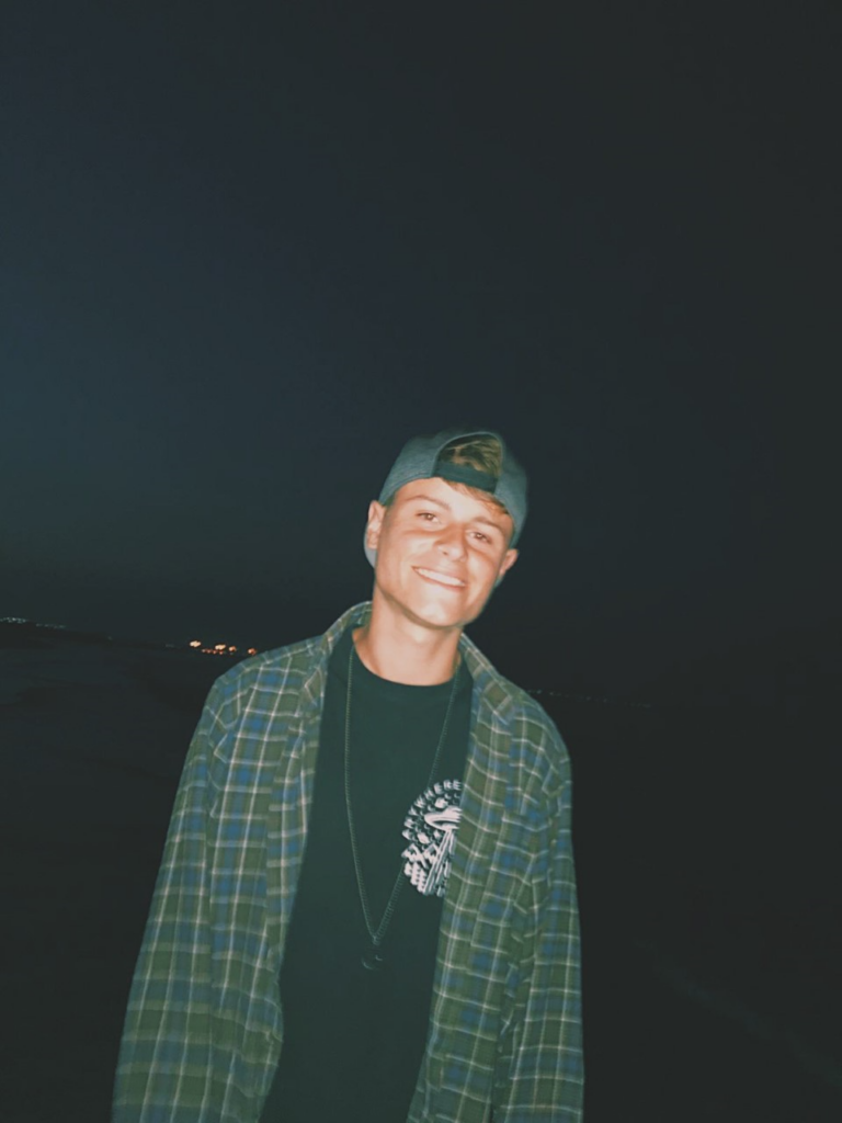 Picture of Matt with a dark background, wearing a cap backwards, a checked shirt and black t-shirt.