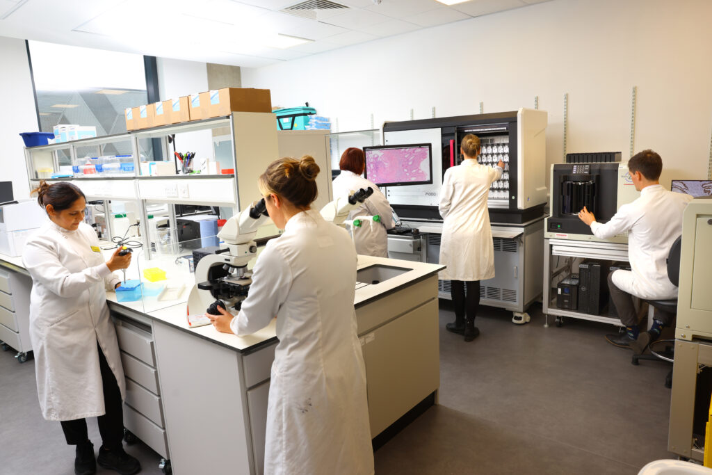 A team of five scientists in white coats works on equipment at work stations in a lab.