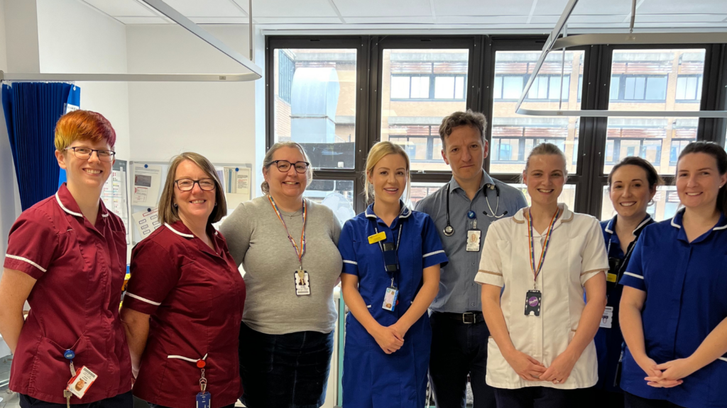 Nurses and doctors stand in a group on a hospital ward. Two of the nurses are in burgundy uniform, one in white and the other three in royal blue. In the middle of the photo is Dr Gherorghiu, a stroke doctor. 